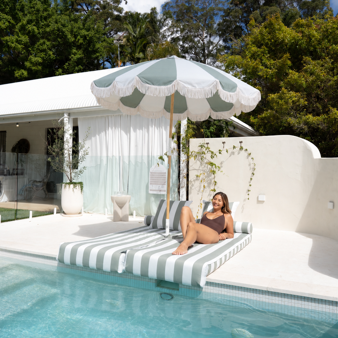 A woman relaxes on a striped poolside lounger under the Mentari Market Umbrella in Sage and oat